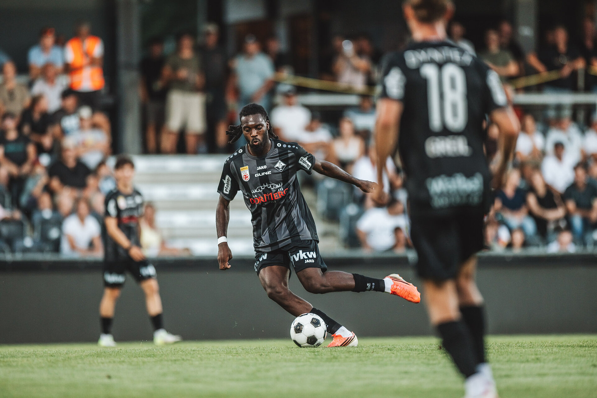 Fußballspieler mit Dreadlocks spielt Ball auf Rasen vor Zuschauern in einem Stadion