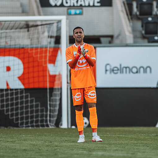 Torhüter Patrick Pentz von Austria Wien klatscht in orangefarbener Spielkleidung auf dem Rasen.
