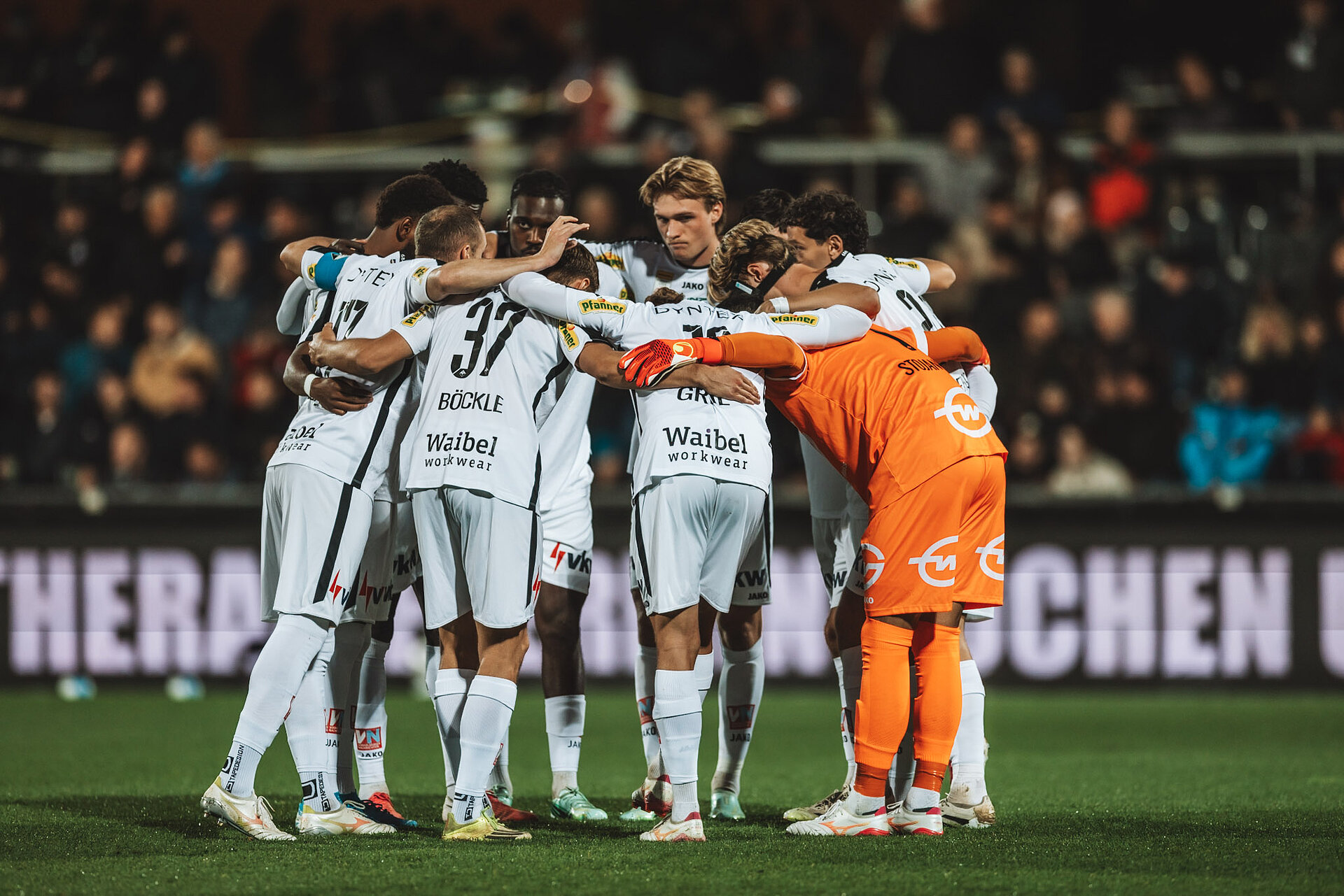 Fußballmannschaft im Kreis stehend mit Torwart im orangen Trikot auf einem Rasenplatz