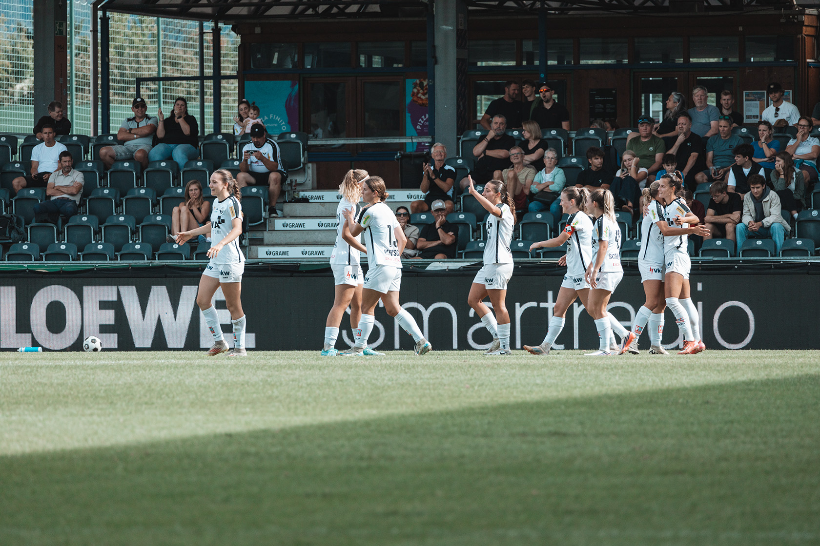 Fußballerinnen in weißen Trikots jubeln auf einem Rasen vor Zuschauern im Stadion.